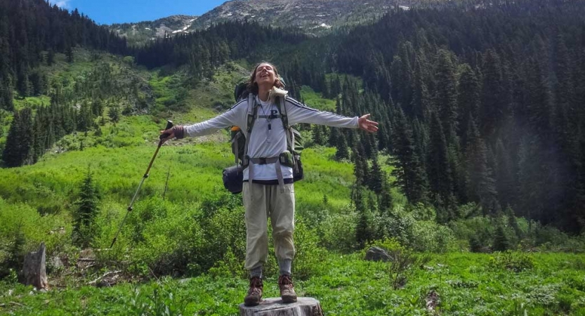 A person standing on a stump spreads their arms out wide. Behind them is a green valley.